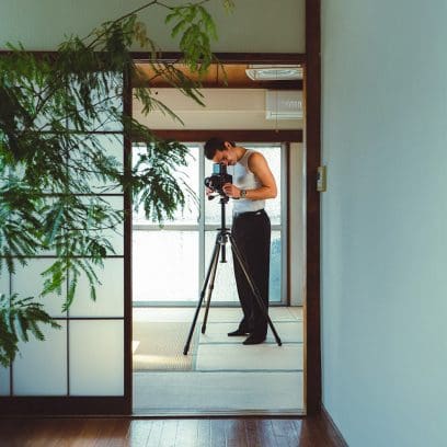 Um homem está junto a um tripé com uma câmara SLR, vestindo uma camisa branca, calças pretas e o relógio digital CASIO Timeless Collection AE-1200WHD-1AV.