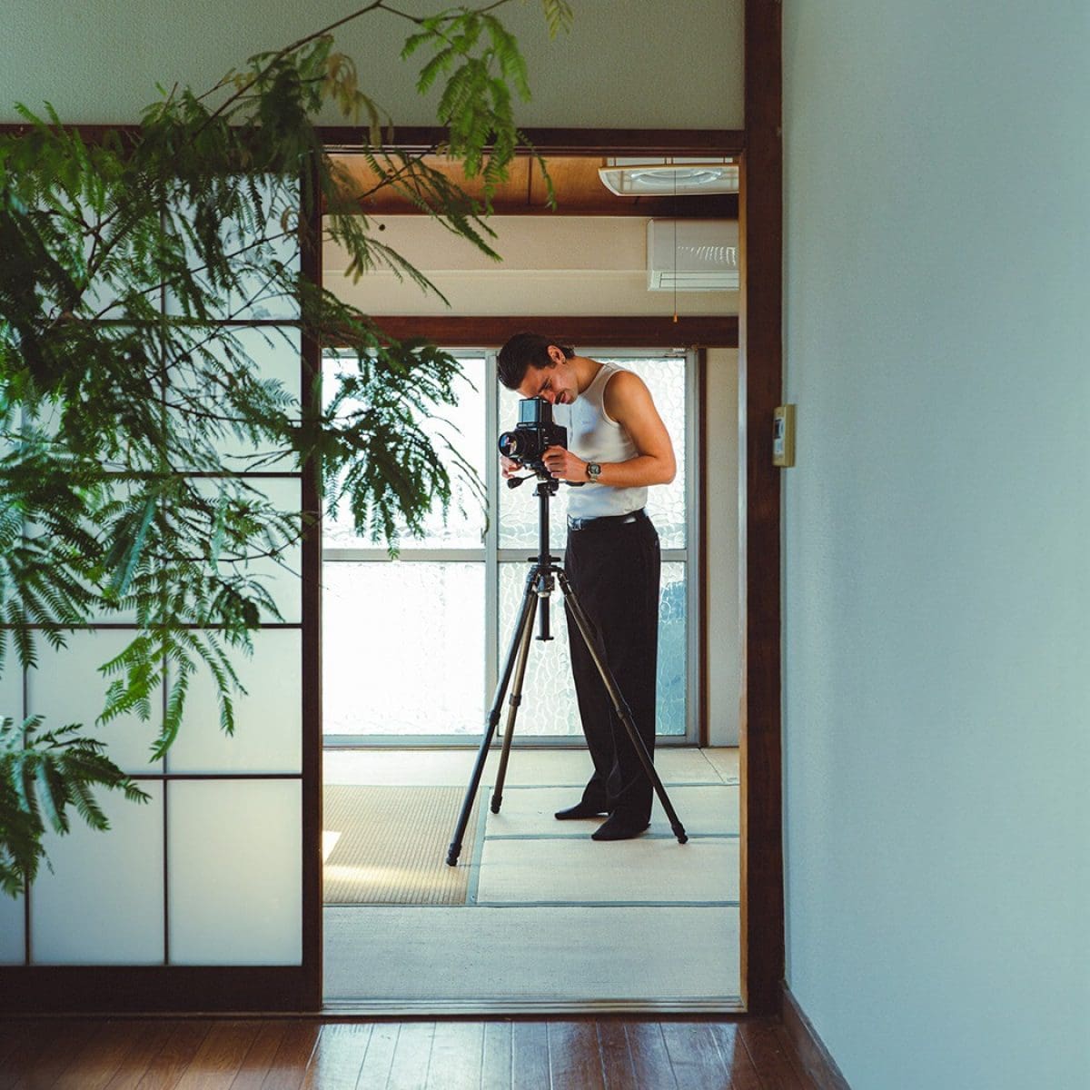Um homem está junto a um tripé com uma câmara SLR, vestindo uma camisa branca, calças pretas e o relógio digital CASIO Timeless Collection AE-1200WHD-1AV.