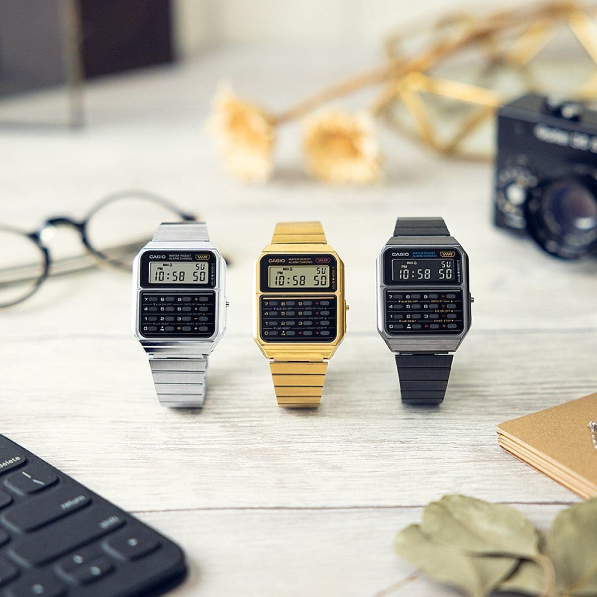 Três relógios da Coleção Vintage da CASIO: CA-500 com bracelete dourada, prateada e preta numa secretária ao lado de uma calculadora, óculos e uma máquina fotográfica