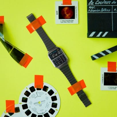 O relógio de pulso CA-500WEGG-1B em cinzento com função de calculadora, com fita adesiva cor de laranja colada num fundo amarelo, ao lado de Polaroids e de uma aba de máquina fotográfica