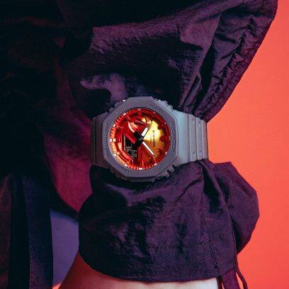O relógio de pulso GA-2100FL-8A da G-SHOCK com um mostrador cor de laranja no pulso. Por baixo, vê-se uma tampa roxa.