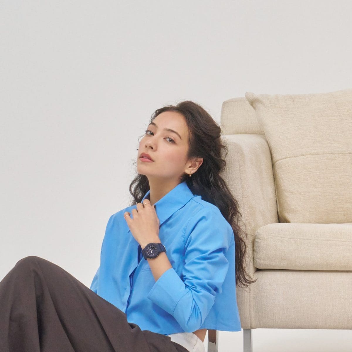 Uma mulher vestindo uma blusa azul clara encostada em um sofá e usa o GMA-P2100M-2A azul no pulso