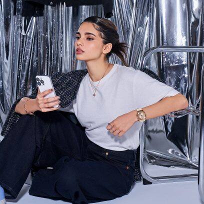 Uma mulher de cabelos castanhos e camisa cinza segura um smartphone na mão e senta no chão, encostada em uma cadeira de metal. No outro pulso ela usa o G-SHOCK GM-S2110PG-4A