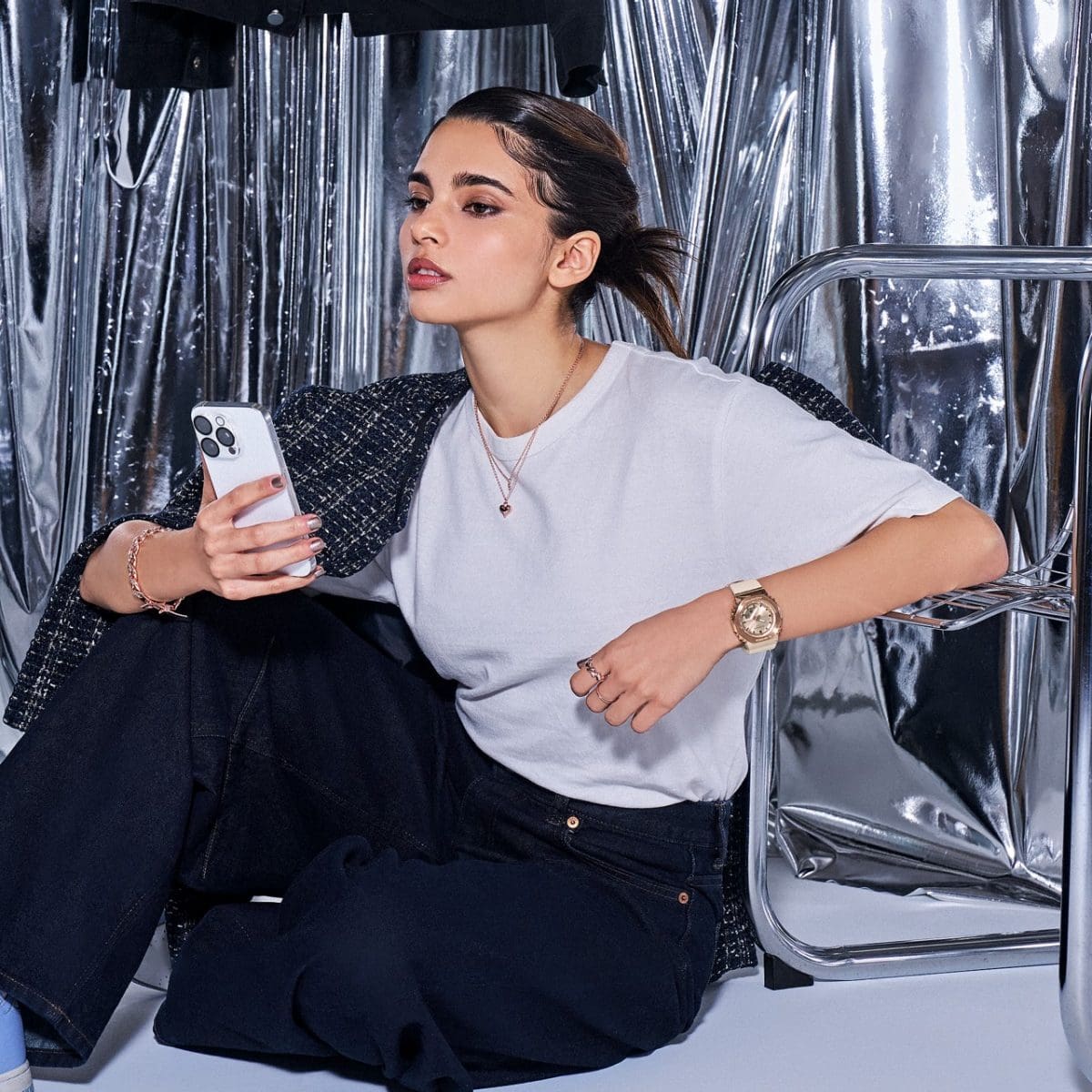 Uma mulher de cabelos castanhos e camisa cinza segura um smartphone na mão e senta no chão, encostada em uma cadeira de metal. No outro pulso ela usa o G-SHOCK GM-S2110PG-4A