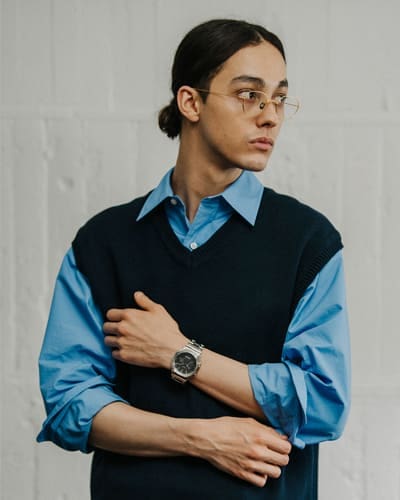 Ein junger Mann mit Pferdeschwanz, Brille, blauem Hemd und Pullunder trägt an seinem linken Handgelenk die silberne Casio G-Shock GM-B2100D Edelstahluhr mit schwarzem Zifferblatt 