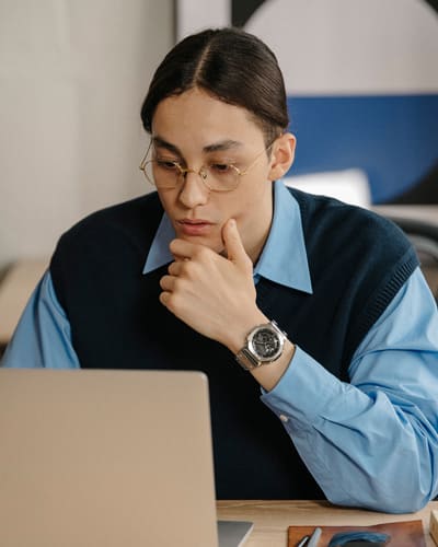 Eine junge Person mit Brille, blauem Hemd und Pullunder sitzt vor einem Laptop, am linken Handgelenk trägt sie die silberne Casio G-Shock GM-B2100D Edelstahluhr mit schwarzem Zifferblatt