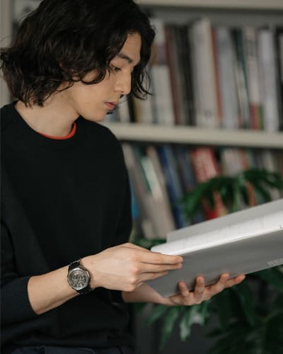 Ein junger Mann mit schulterlangen, dunkeln Haaren steht vor einem Regal und liest in einem Buch, an seinem linken Handgelenk trägt er die silberne Casio G-Shock GM-B2100D Vollmetalluhr
