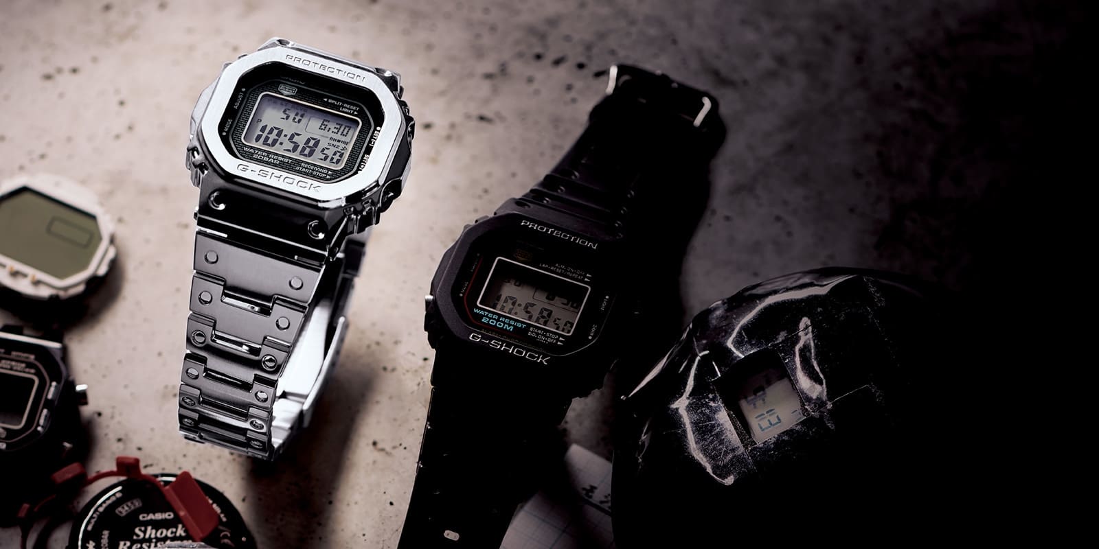 Eine silberfarbene Vollmetalluhr von Casio G-Shock liegt neben einer schwarzen G-Shock Digitaluhr auf Betonboden, drumherum liegen Gehäuseteile anderer Armbanduhren