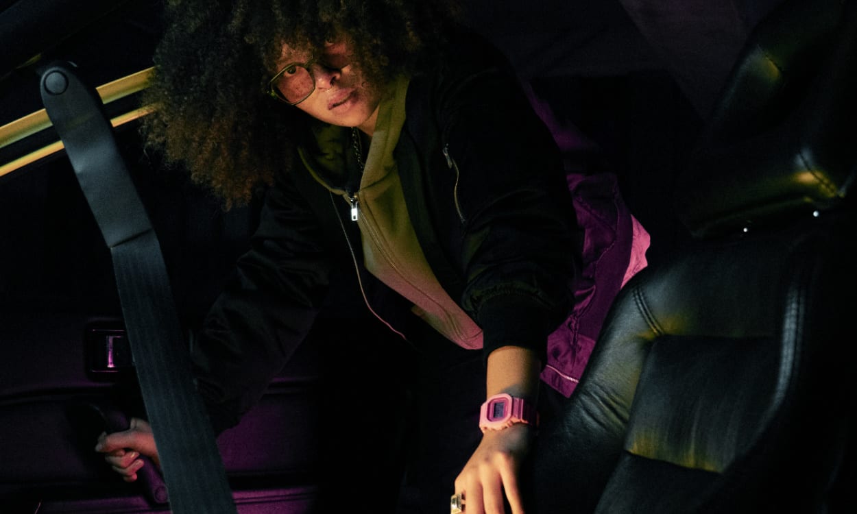 A woman with an afro, a black jacket and the pink G-SHOCK GMD-S5610 leaning into the passenger seat of a car.