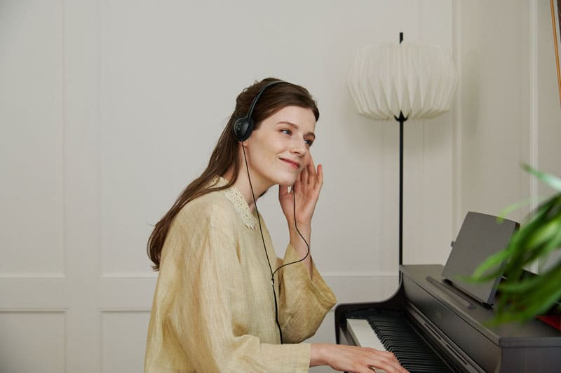 Eine Frau mit langen, offenen, dunkelbraunen Haaren trägt eine helle Bluse und sitzt an einem dunkeln Digitalpiano AP-300 von CASIO. Sie trägt Kopfhörer und spielt Klavier