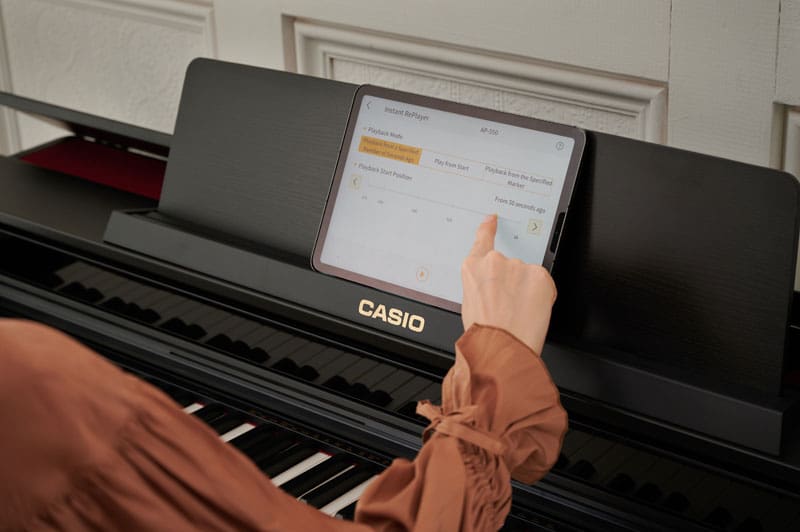 Eine Frau sitzt an einem dunkeln Digitalpiano von CASIO, im Notenständer des Klaviers steht ein Tablet mit der "CASIO MUSIC SPACE"-App