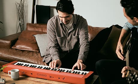 Ein Mann sitzt auf einem braunen Ledersofa. Auf dem Wohnzimmertisch vor ihm steht ein rotes Digitalpiano CT-S1RD auf dem er klavierspielt.