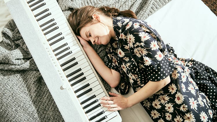 Eine glücklich lächelnde junge Frau liegt auf einer grauen Strickdecke neben einem weißen CT-S1WE und berührt die Tasten des Digitalpianos