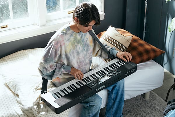 Ein junger Mann in einem hellen Batikpullover sitzt auf einem Bett und spielt auf einem Casiotone CT-S500, das auf seinem Schoß liegt