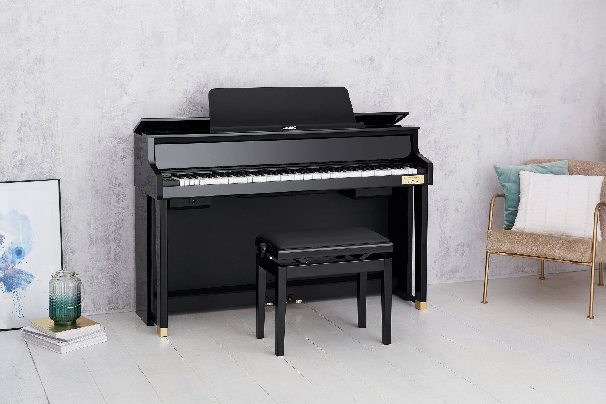 Das glänzend schwarze Celviano Grand Hybrid Digitalpiano GP-510BP steht an einer grauen Zimmerwand, links daneben lehnt ein Bild an der Wand, rechts steht ein Korbstuhl mit Kissen 