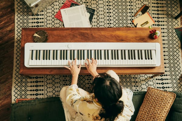 Blick von oben auf eine Person, die auf einem Sofa sitzt und auf einem weißen Privia PX-S1100WE-Digitalpiano klavierspielt, das auf einem Wohnzimmertisch aus Holz vor der Person liegt