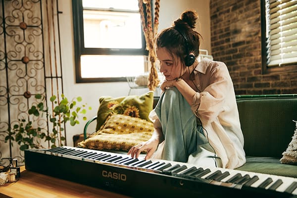 Eine junge, Kopfhörer tragende Frau sitzt auf einem Sofa und spielt auf dem schwarzen Privia PX-S1100BK Digitalpiano, das vor ihr auf dem Wohnzimmertisch liegt