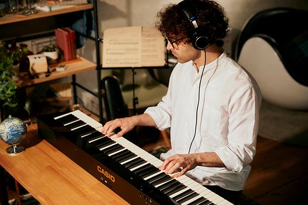 Seitlicher Blick von vorne auf eine Kopfhörer tragende Person, die an einem Privia PX-S5000-Digitalpiano klavierspielt, das auf einem Tisch vor der Person liegt