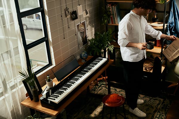 Ein junger Mann in weißem Hemd steht in einem kleinen Wohnraum und schaut auf ein Notenblatt, das auf einem Notenständer liegt. Hinter ihm steht vor einem Fenster ein PX-S5000 Digitalpiano auf einem Schreibtisch.