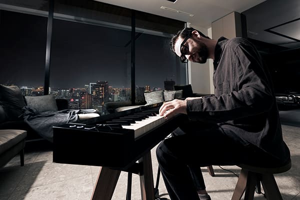 Ein Mann spielt in gedimmtem Licht auf dem PX-S7000BK Digitalpiano, während durch Panoramafenster eine nächtliche Großstadt zu sehen ist
