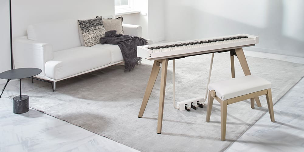 Ein heller Wohnraum mit großen Fenstertüren zum Meer, in der Mitte des Raumes steht ein Privia PX-S7000HM Digitalpiano mit passendem Klavierhocker. Text ergänzt das Bild: „Style, Reimagined – PX-S7000 – Privia“. Die Seite verlinkt zur Privia-Info-Seite.