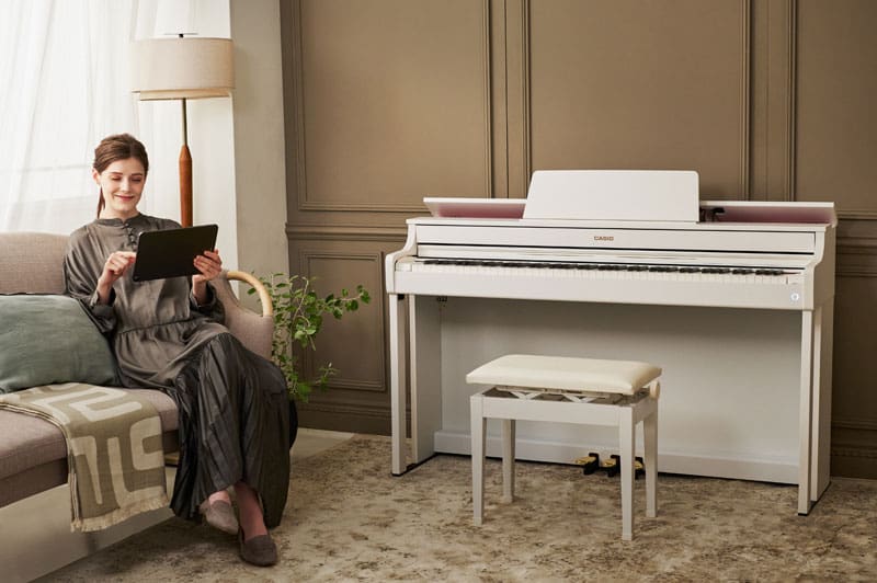 A woman in a long dark grey dress is sitting on a sofa in a living room, smiling contentedly as she looks at a tablet. On the wall next to her is a white AP-300 digital piano with a matching piano bench.