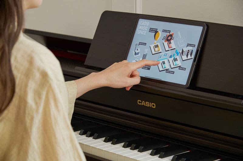 A woman with long dark brown hair and a light blouse is sitting at a dark AP-300 digital piano and working on a tablet with the ‘CASIO MUSIC SPACE’ app.