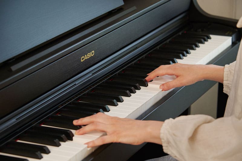 View of the keys of a black CASIO AP-300 digital piano and two women's hands touching them to play the piano