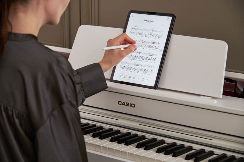 A woman wearing a dark grey blouse sits at a white CASIO AP-300 digital piano and works with a digital sheet of music on a tablet with her left hand. 