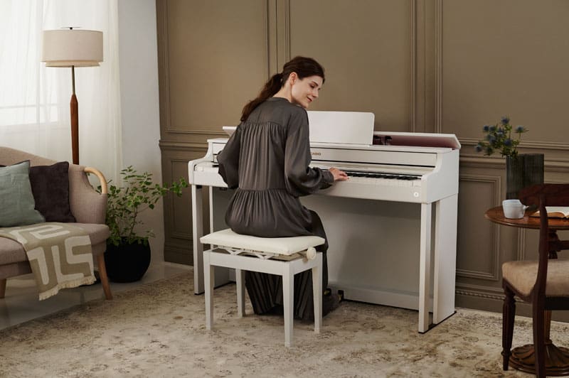 A woman with long dark brown hair and wearing a dark grey dress is sitting in a living room at a white digital piano AP-300 from CASIO and plays the piano