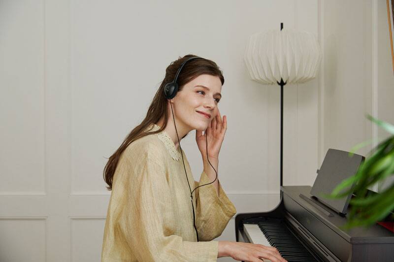 A woman with long, open, dark brown hair, wearing a light blouse and sitting at a dark digital piano AP-300 from CASIO. She is wearing headphones and playing the piano.