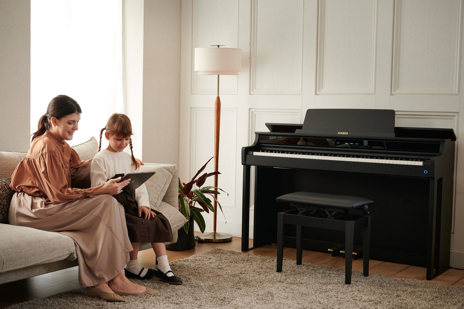 A woman is sitting on a sofa with a girl. They are looking at something on a tablet together. On the wall of the comfortably furnished room is a black CELVIANO AP-550BK digital piano with a matching piano bench