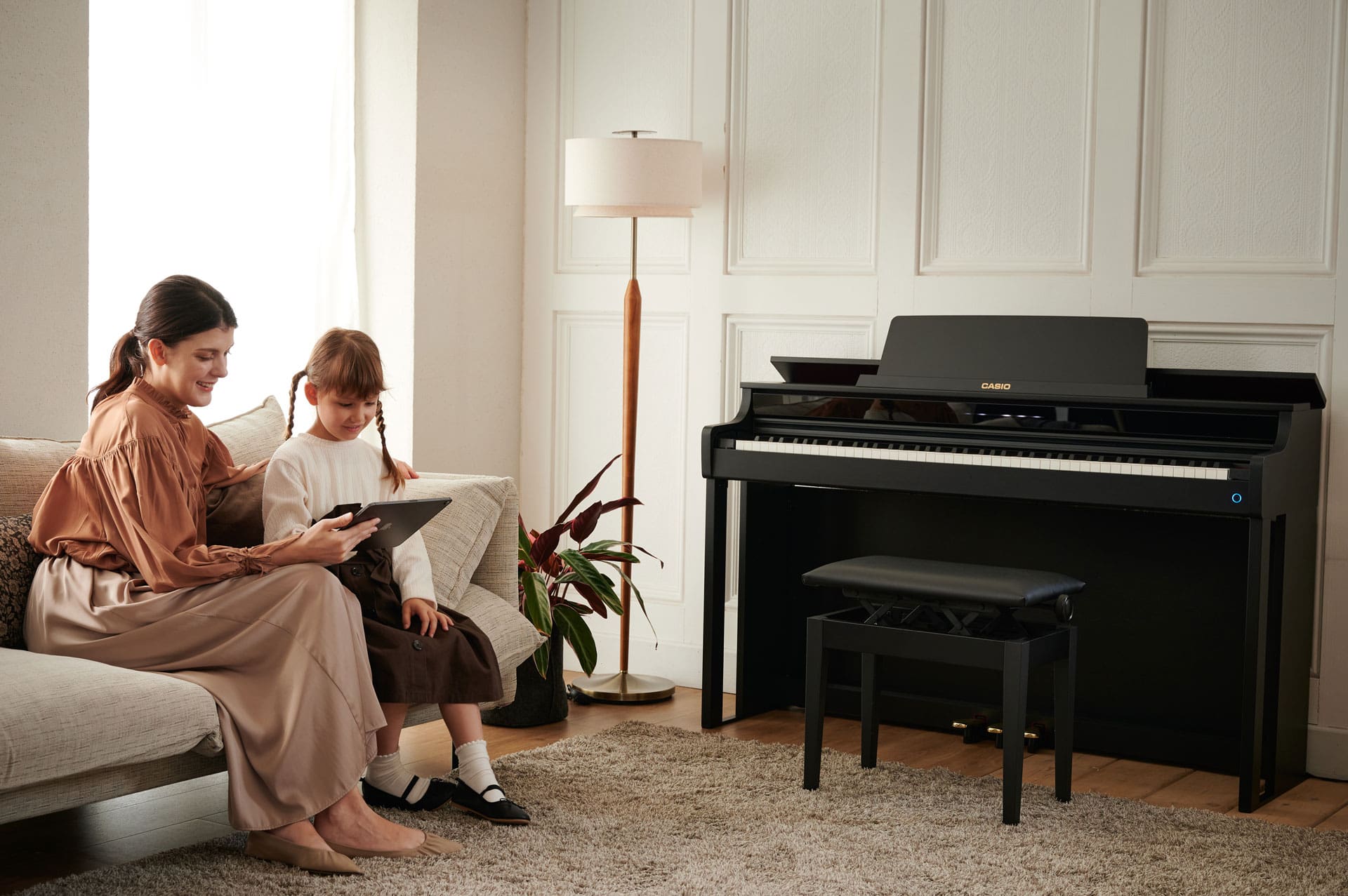 A woman is sitting on a sofa with a girl. They are looking at something on a tablet together. On the wall of the comfortably furnished room is a black CELVIANO AP-550BK digital piano with a matching piano bench
