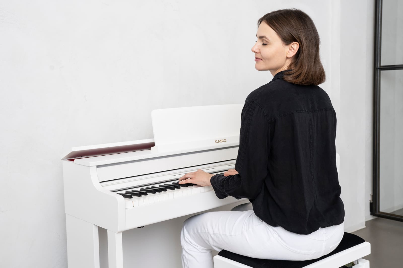 A woman sits in a bright room at a white CELVIANO AP-550WE digital piano, completely absorbed in her piano playing.