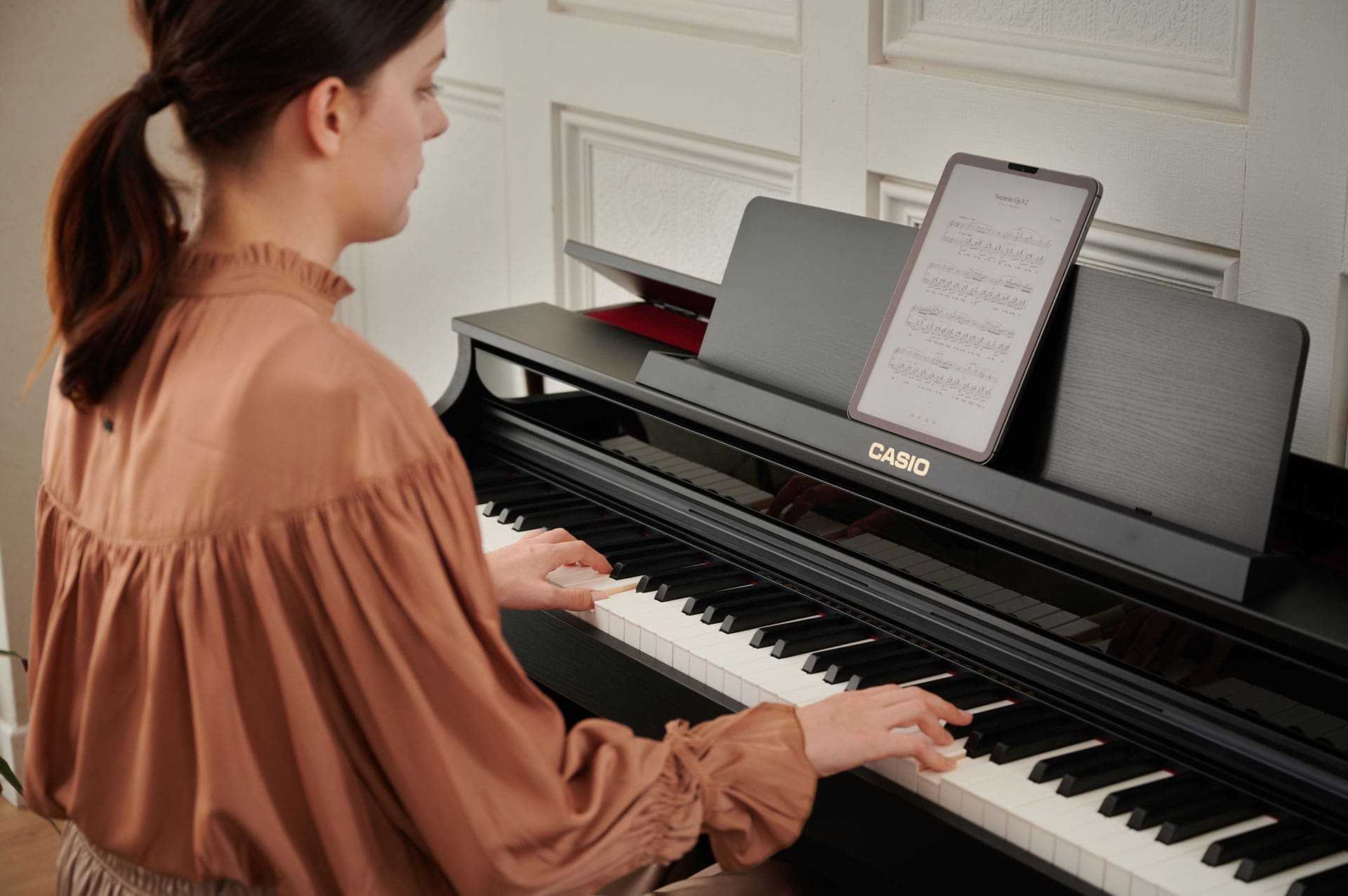 A woman sits at the black CELVIANO AP-550BK and plays the piano. A tablet with digital sheet music is placed on the music stand of the digital piano. 
