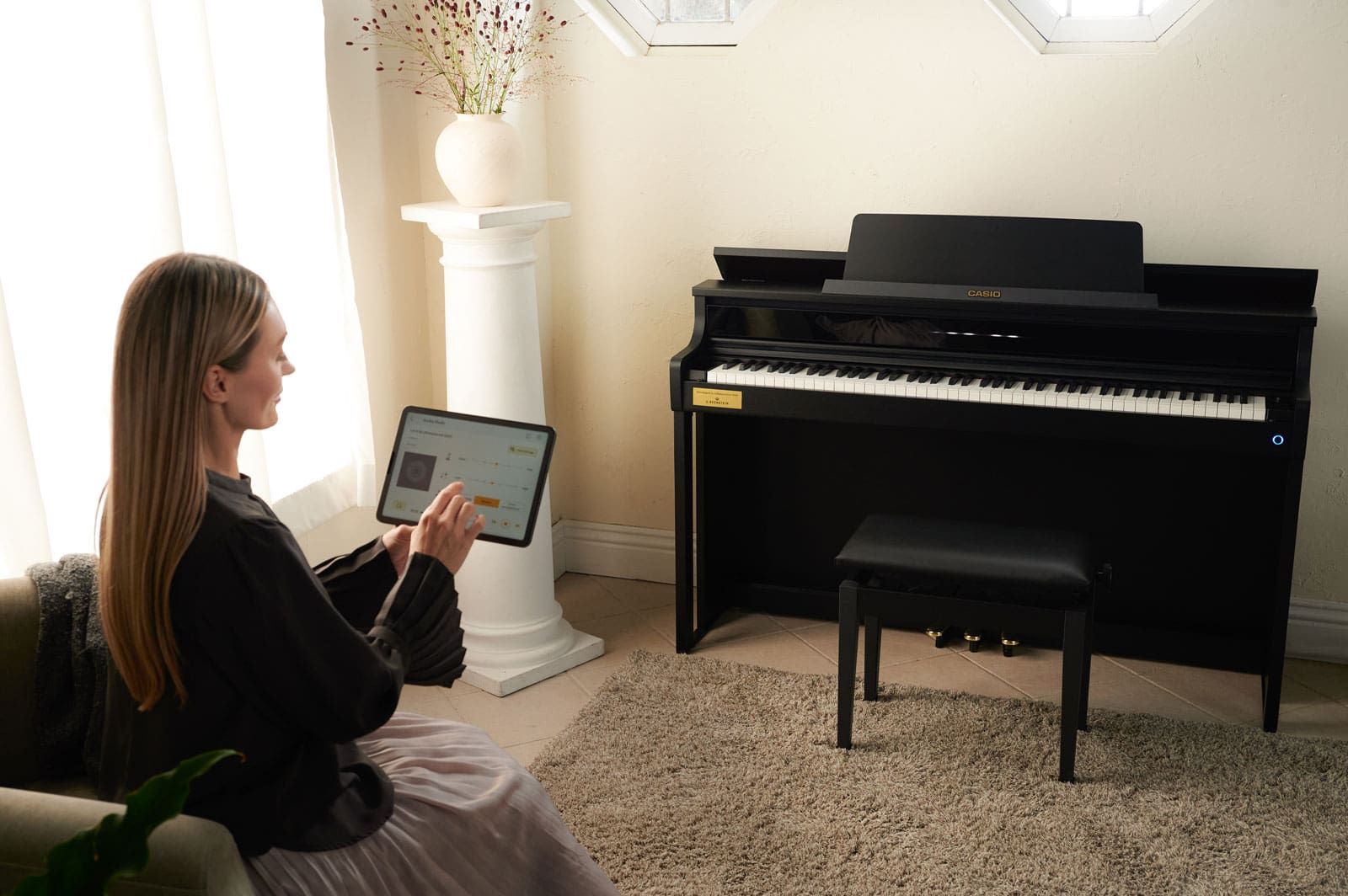 The CASIO AP-750 digital piano stands against the wall of a room, a woman sits on an armchair nearby and works with a tablet in the CASIO MUSIC SPACE app