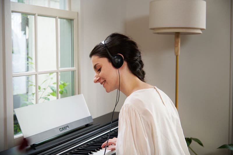 A woman wearing headphones sits smiling at a dark AP-S200 digital piano and plays the piano