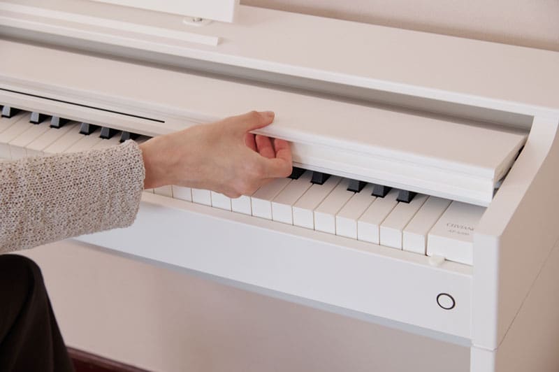 A hand reaches for the half-opened keyboard cover of a white AP-S200WE digital piano