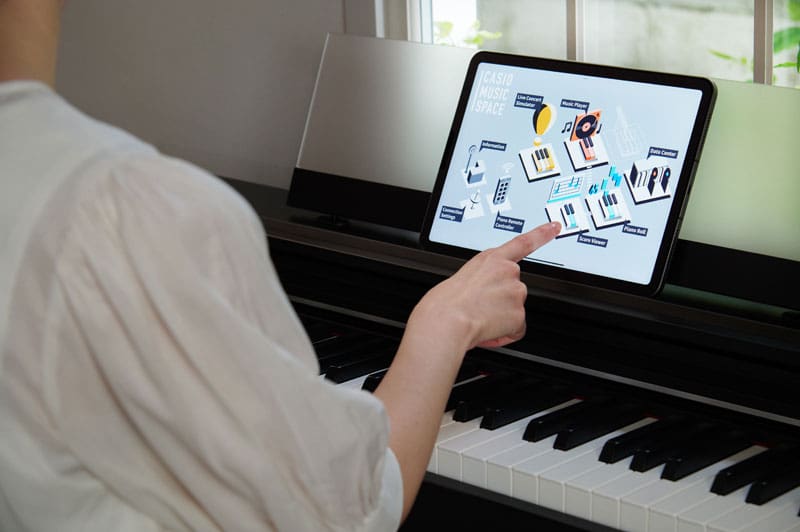 A hand touches a tablet placed in the music stand of a dark AP-S200 digital piano to operate the ‘CASIO MUSIC SPACE’ app.
