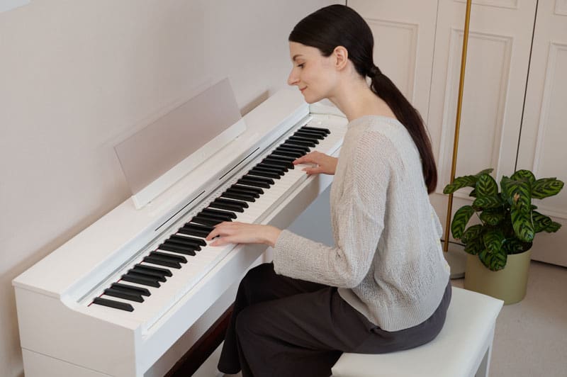A woman is sitting smiling at a white Celviano digital piano AP-S200WE and playing the piano