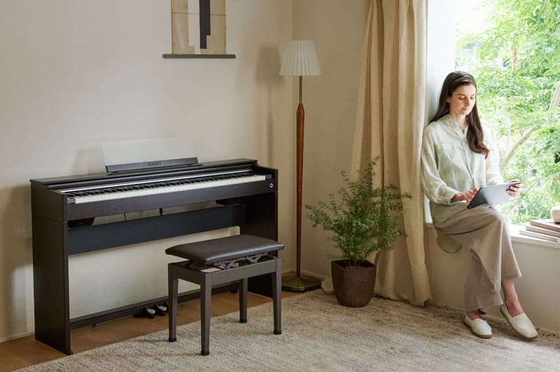A young woman sits on a windowsill and looks at a tablet, on the wall to her right is a brown AP-S200BN digital piano
