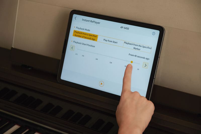 A hand touches a tablet placed in the music holder of a dark AP-S450 digital piano to operate the ‘CASIO MUSIC SPACE’ app