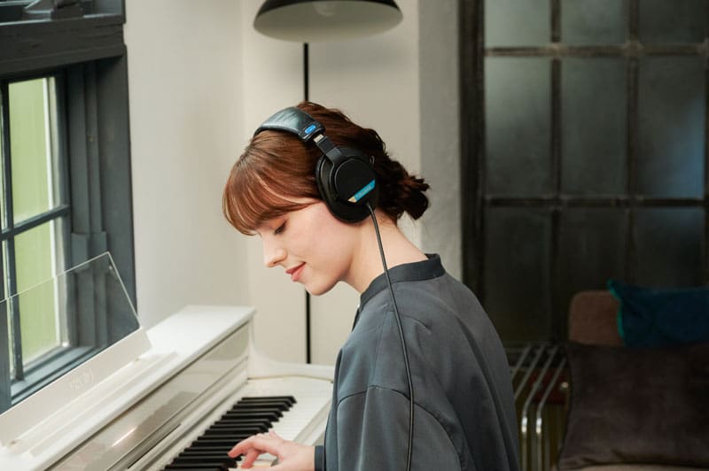 A young woman wearing headphones sits smiling at a white AP-S450 digital piano and plays the piano