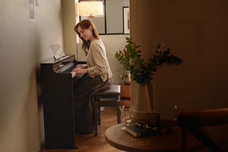 A woman is sitting on a piano bench in a small, warmly lit room, playing the AP-S450 digital piano