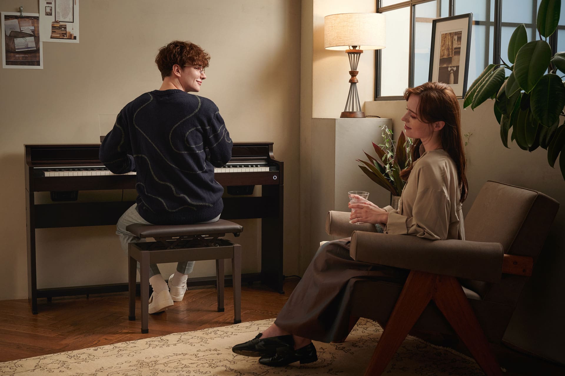 A young man is playing the AP-S450 digital piano in a warmly lit living room, looking over his right shoulder at a woman sitting in an armchair behind him, listening devoutly