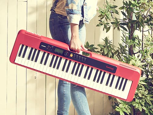 A person is shown in a medium shot holding the red Casiotone CT-S200 keyboard with handle in their hand.