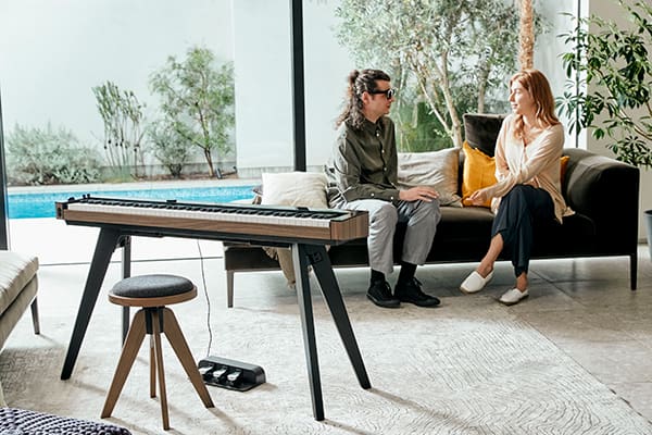The PX-S6000 digital piano is placed on the CS-90P piano stand in a living room. In the background, a man and a woman are sitting on a sofa