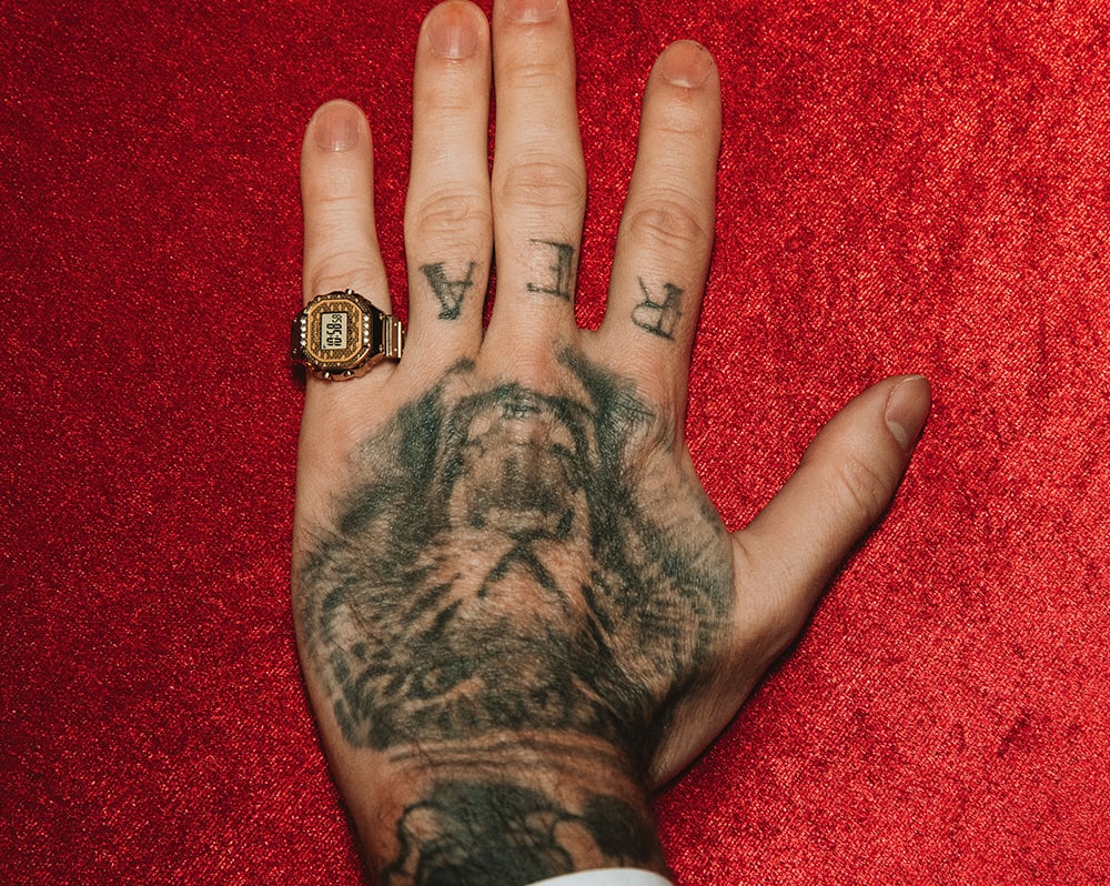 A man's hand wearing CRW-001JB-9 on the pinky finger, on a red background