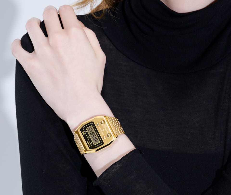 Una persona vestida completamente de negro se sujeta el hombro derecho con la mano izquierda, en la muñeca: el reloj dorado A1100G-5 de la colección vintage de Casio.