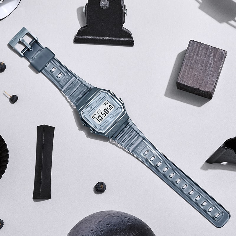 El F-91WS-8 en gris transparente sobre una mesa junto a un clip negro y un trozo de madera gris.